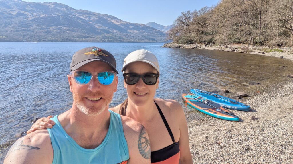 Rob & Julia standing on a beach on the east bank of Loch Lomond after paddleboarding.
